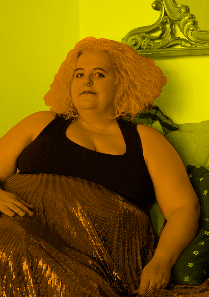 Elisa Manici sits down on a couch. They wear a black top and sequined skirt. The image, framed in black and resting on a red background, is filtered in yellow and orange, which alternates in flashes on their silhouette and the background.